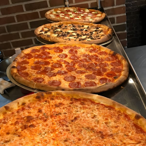 Four large, freshly baked pizzas with various toppings, including pepperoni, on metal trays.